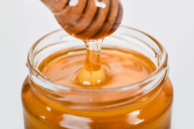 Close-up of honey flowing from a wooden dipper into a glass jar on a white background.